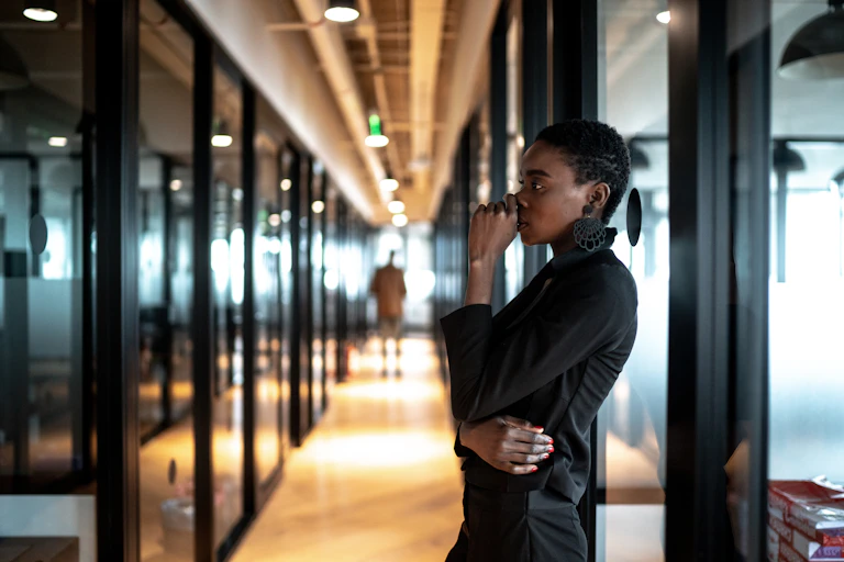 A woman leans up against an office wall and holds her face, looking concerned