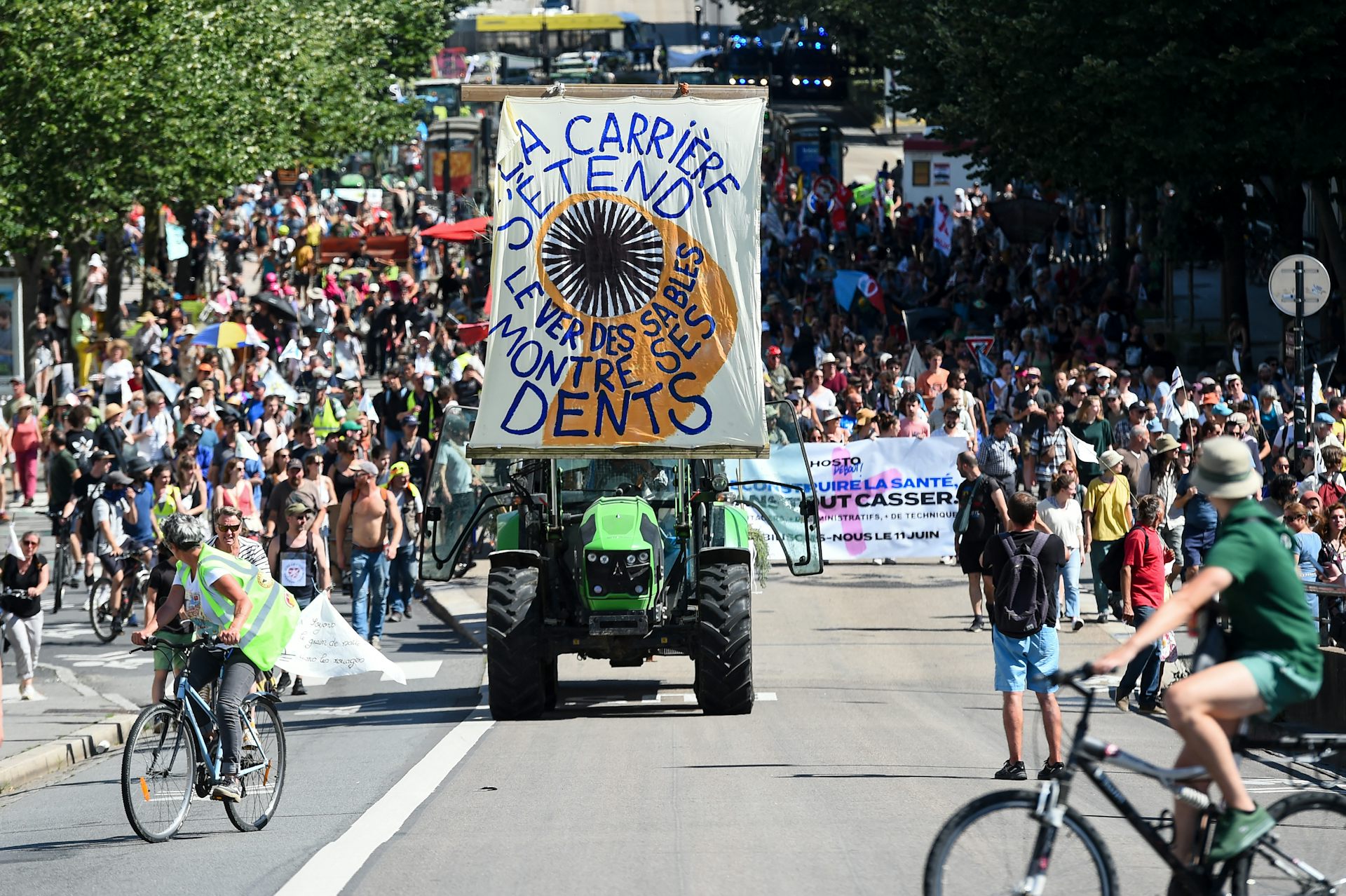 Manifestation avec des piétons, des véols et un tracteur avec une banderole sur on lit « La carrière s'agrandit, le ver des sables montre les dents »