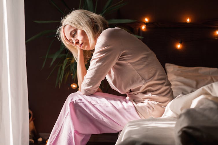 A woman sits on the edge of a bed with her head in her hand.