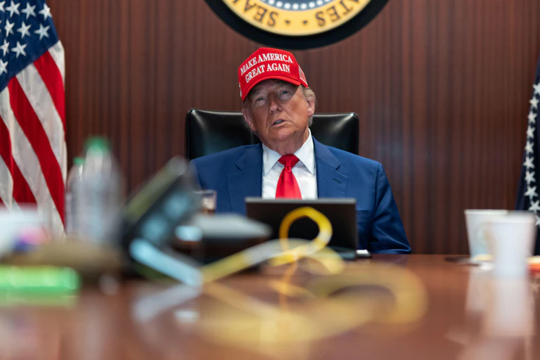 US president, Donald Trump sits behind a desk wearing a Maga hat and watching a screen.