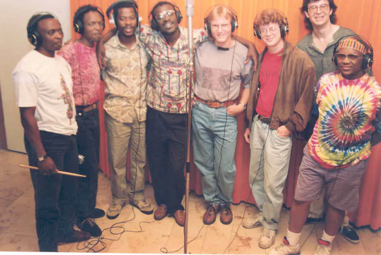 A vintage colour photo of eight musicians in a row in a recording studio.