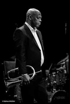 A black and white photo of an African man in a suit in profile, a trumpet in his hand, his eyes closed and head lowered.