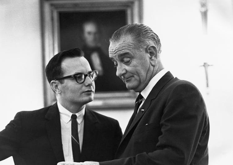 LBJ speaks with a young man with dark-rimmed glasses who is wearing a 1960s-style suit and skinny tie.