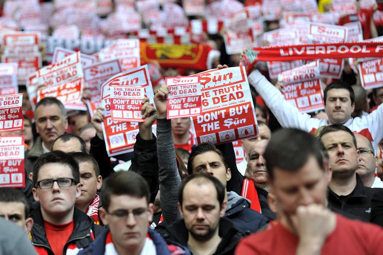 A stadium crowd holding up red and white signs that read 'the truth 96 dead, don't buy the Sun'
