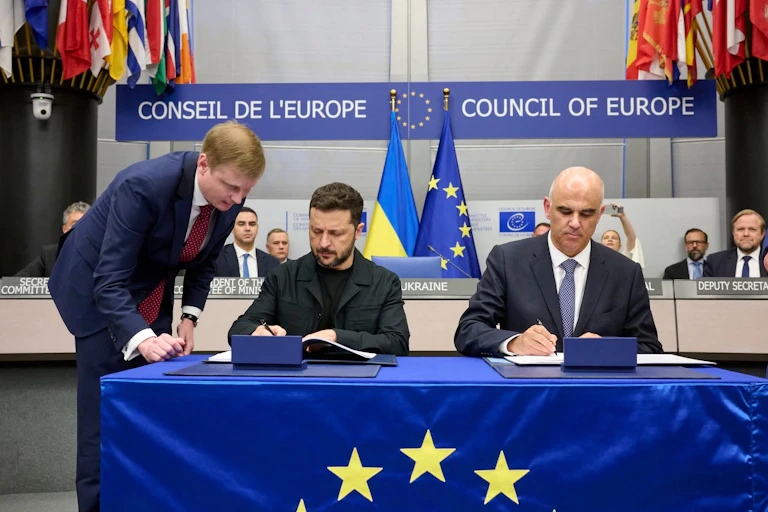 Ukrainian president Volodymyr Zelensky sits at a table next to Council of Europe secretary general, Alain Berset, signing a document.