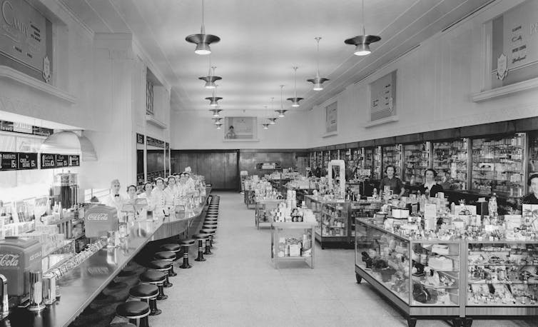 Black and white photo of the interior of a drug store, with various health remedies sold on the right side, and a soda fountain with stools on the left.