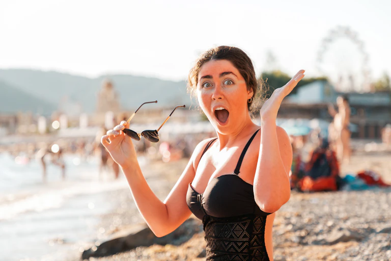 Woman holds sunglasses and screams from sunburn. Redness on the skin of the face and body. In the background, the beach and the sea.