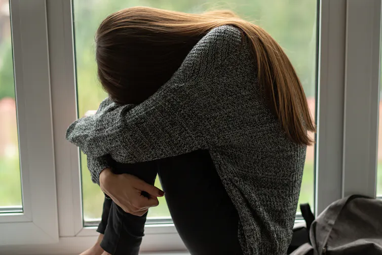 A young **missing word** buries her head between her knees and cries near a window.
