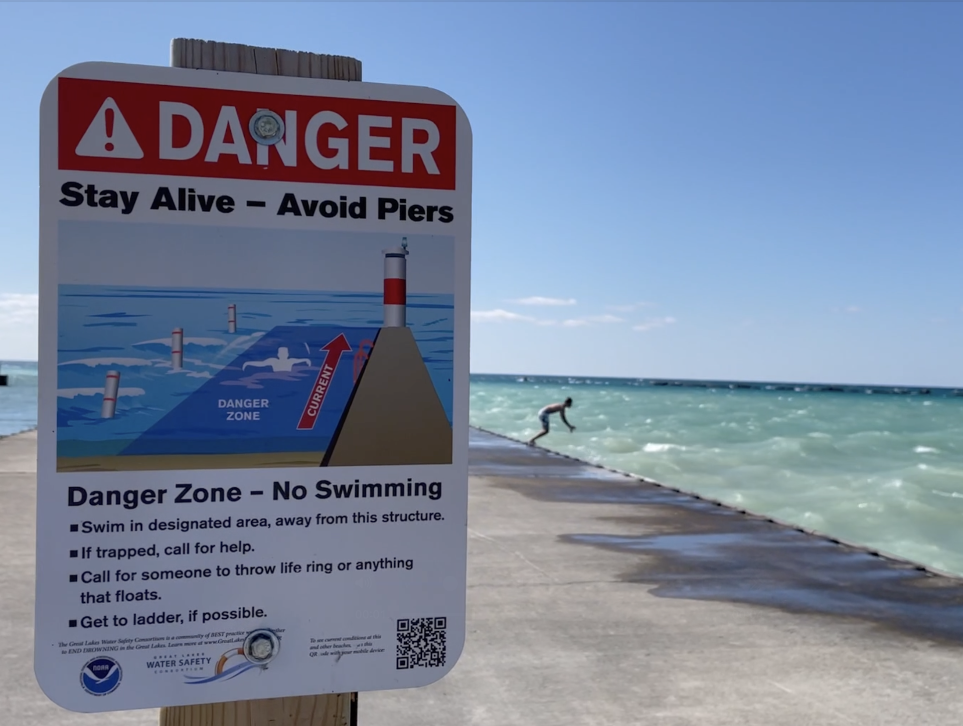 A sign in the foreground warning 'Danger Zone — No Swimming' while a person in the background prepares to jump into the water 