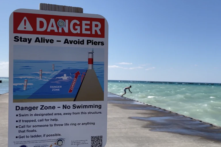 A sign in the foreground warning 'Danger Zone — No Swimming' while a person in the background prepares to jump into the water