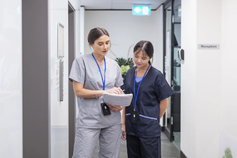 Two women in scrubs look at a notebook in a corridor