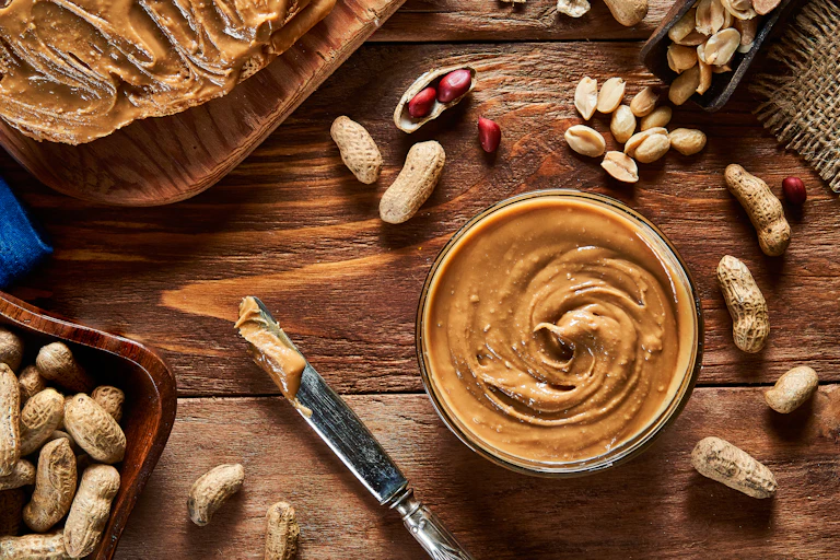 Peanut butter in a jar next to a smeared knife and some peanuts.