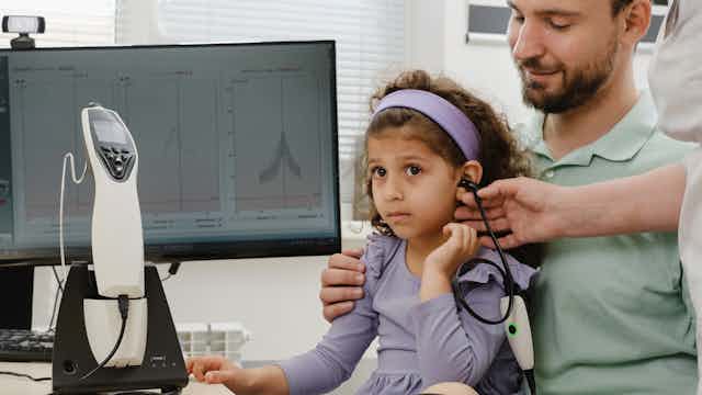 A child sits on her father's lap while undergoing a hearing test at the audiologist.