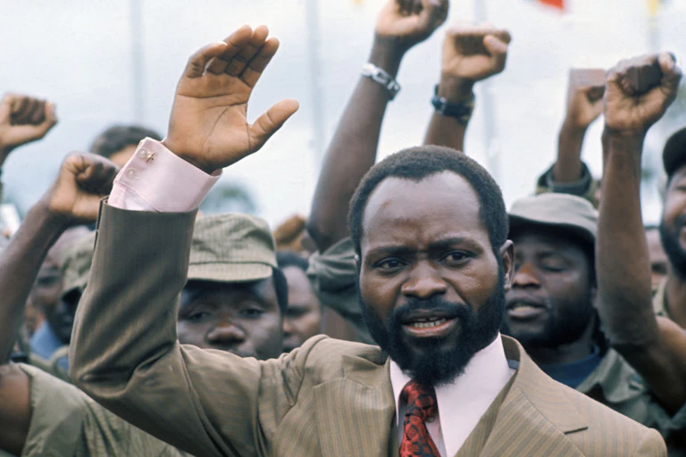 A man in suit and tie, with his hand raised above his head. Behind him other men raise clenched fists.