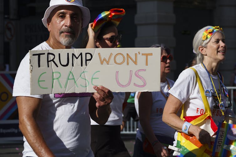 A man dressed in a white hat and tshirt holds a sign that reads 'Trump won't erase us.'