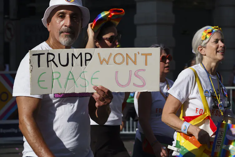 A man dressed in a white hat and tshirt holds a sign that reads 'Trump won't erase us.'