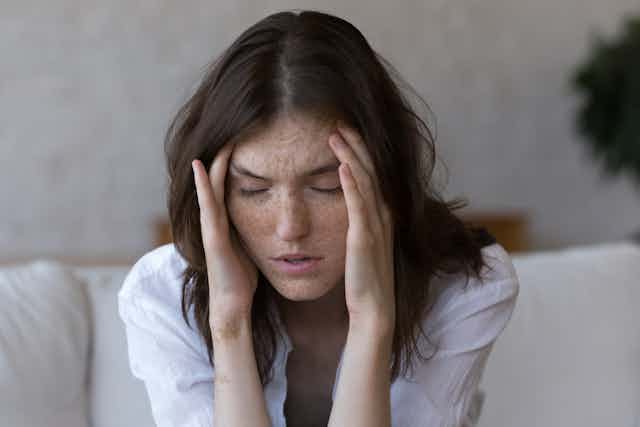 A women with freckles holds her fingers against her temple and closes her eyes. She has the look of someone with a severe headache.