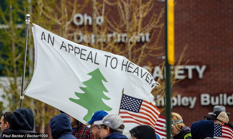 Sebuah bendera putih bertuliskan kata -kata 'sebuah banding ke surga,' yang menampilkan pohon pinus hijau, dengan bendera Amerika ditampilkan di bawahnya.