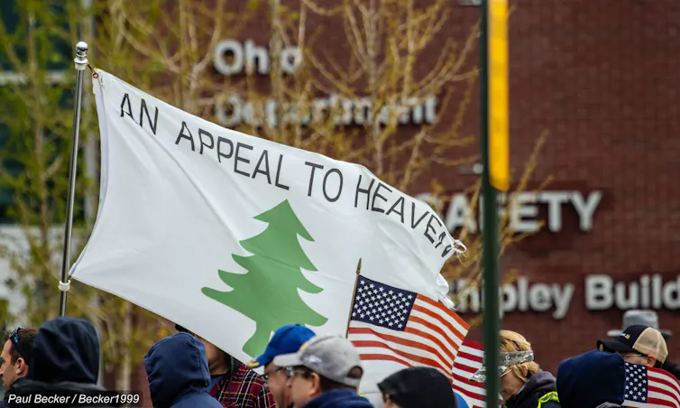 A white flag bearing the words 'An Appeal to Heaven,' featuring a green pine tree, with the American flag displayed beneath it.