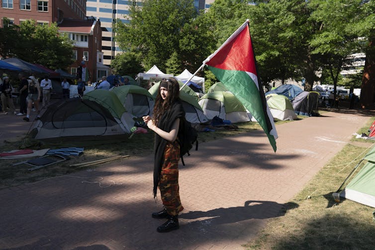 a student holds a green red and white flag on pavement in front of tents on a college campus