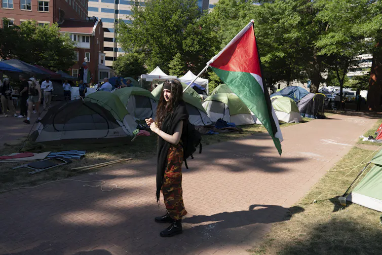 a student holds a green red and white flag on pavement in front of tents on a college campus