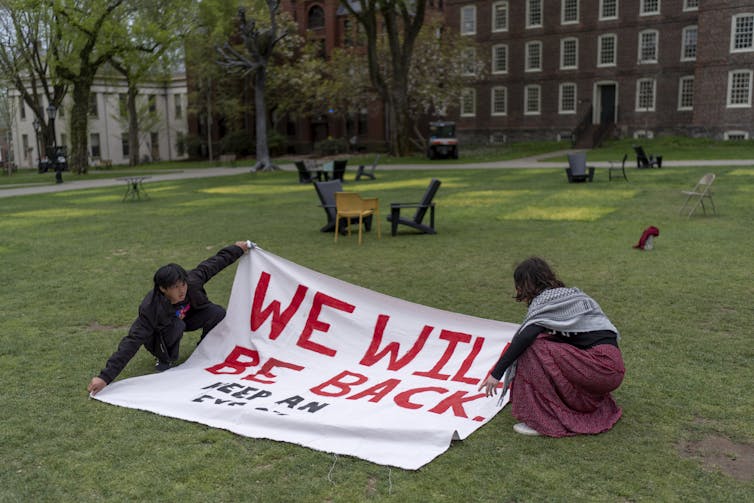 two people hold a sign saying we will be back on a mostly empty college lawn