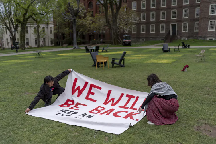 two people hold a sign saying we will be back on a mostly empty college lawn