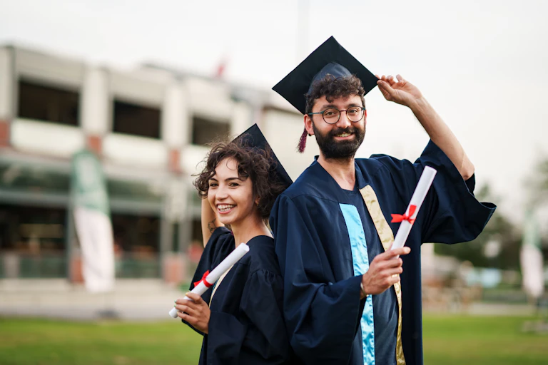 A man and woman pose in black graduation robes and hats on a campus lawn, holding diplomas.
