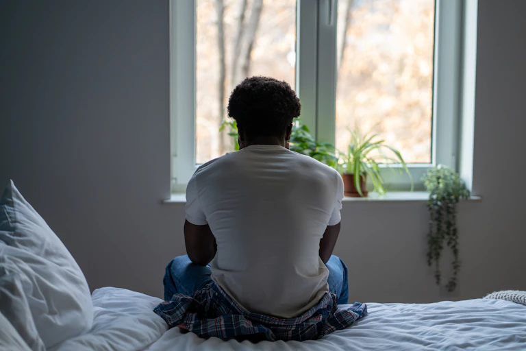 View from behind of man sitting on edge of bed looking out of window lined with houseplants.