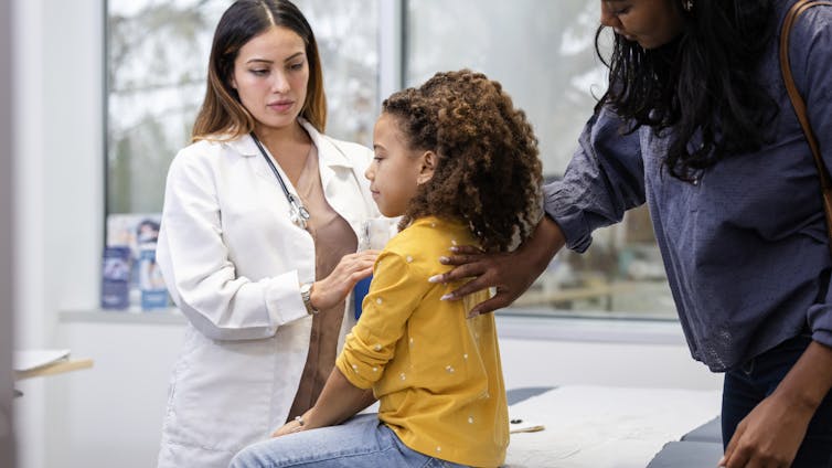 Female doctor with long hair, white coat and stethoscope examines a young child sitting on an exam table, while an adult stands behind the child.