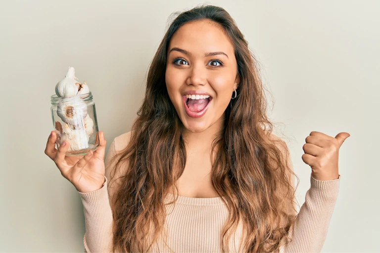 Young woman holds jar of garlic bulbs while giving thumbs up and smiling