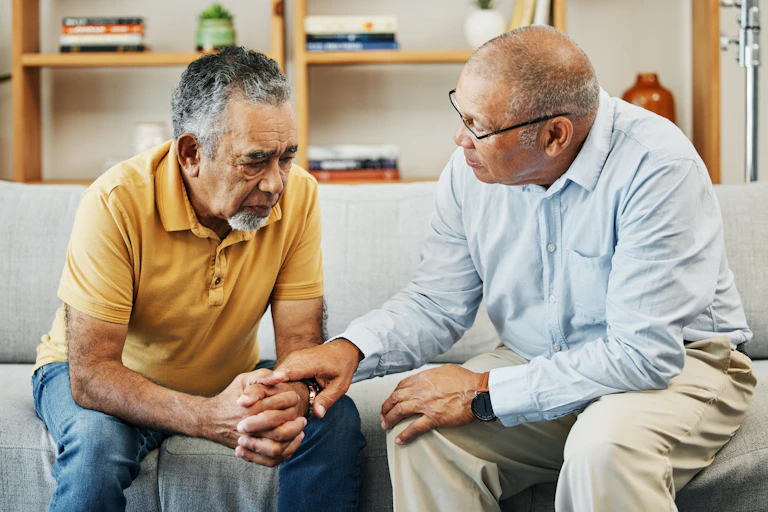 Two men talking on a sofa. One man hold the hand of the other in a supportive gesture.
