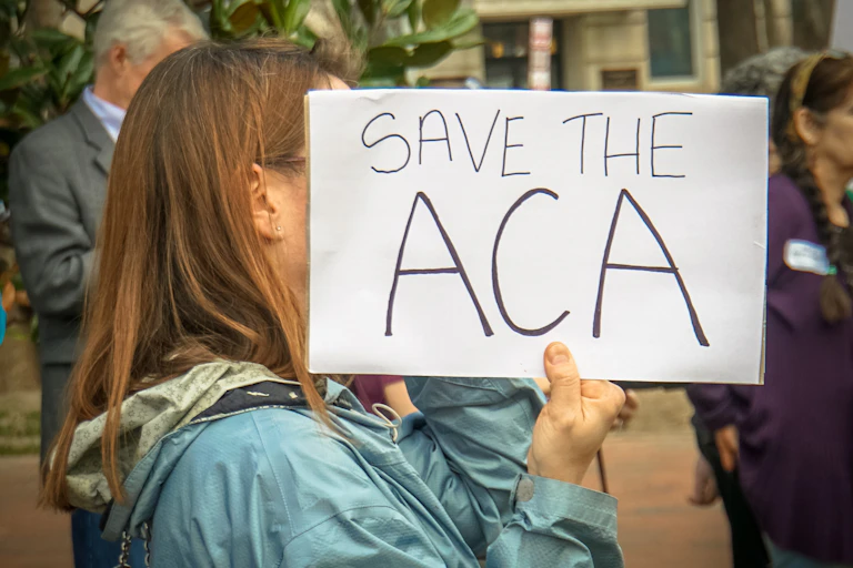 A woman in a light blue raincoat holds a sign covering her face that says SAVE THE ACA.