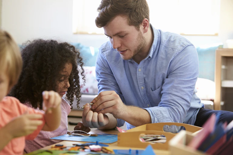 A young man sitting at a table in a childcare centre plays with children