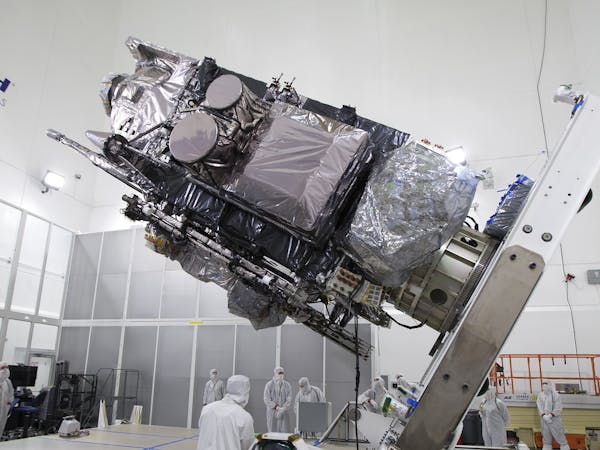 Scientists and engineers in protective white lab clothing use a lift to move a satellite vertical for loading aboard a rocket for launch.