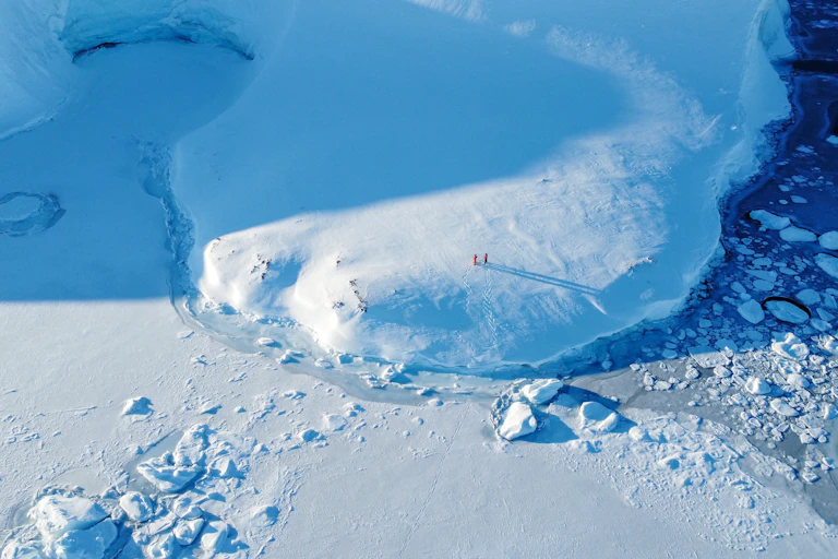 Aerial view of a snowy and icy landscape in Antarctica with two tiny people visible in red snow suits.