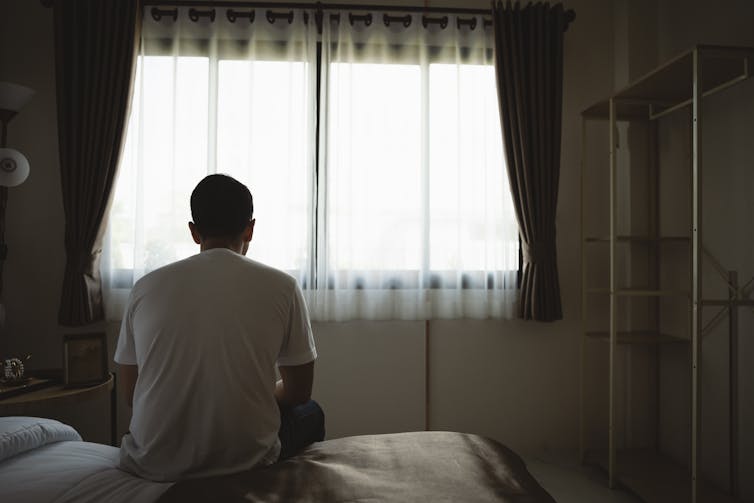 A man sits on his bed, looking out the window.
