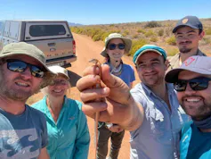 A group of six smiling people stand in the wild and hold up a small orange lizard