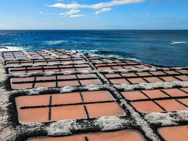 Charcas de unas salinas con el agua rosada y montones de sal entre ellas, con el mar al fondo
