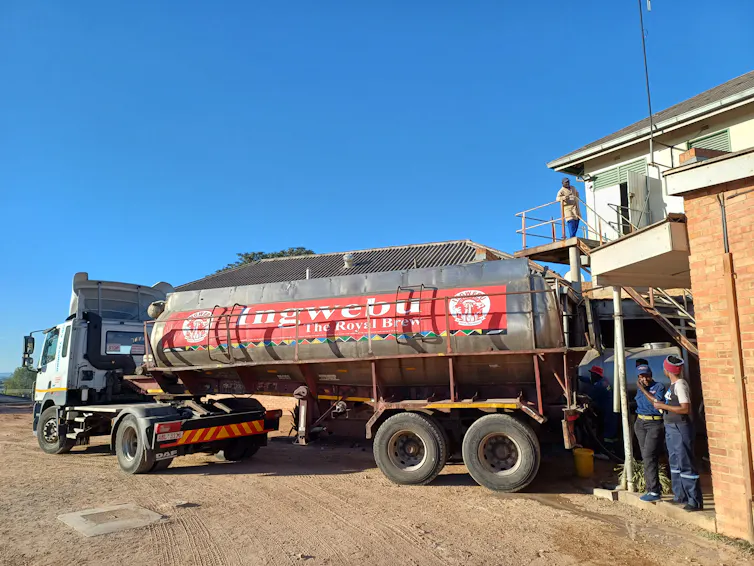 A large tanker with a red logo of a beer company is parked at a building next to some workers in overalls.