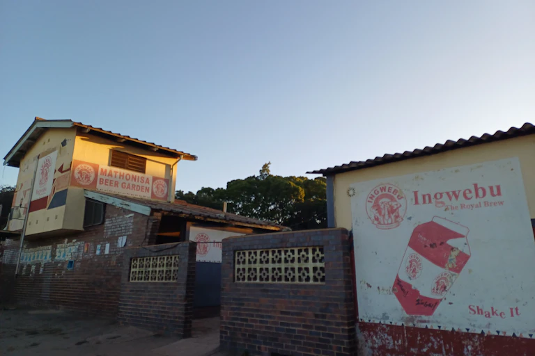An abandoned structure, a large building with "Mathonisa Beer Garden" painted on it and a wall with an advert for "Ingwebu" beer painted on it.