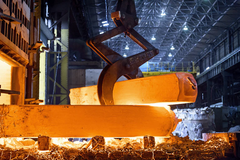 A bright, hot rod of steel in a factory, lifted by a metal arm.