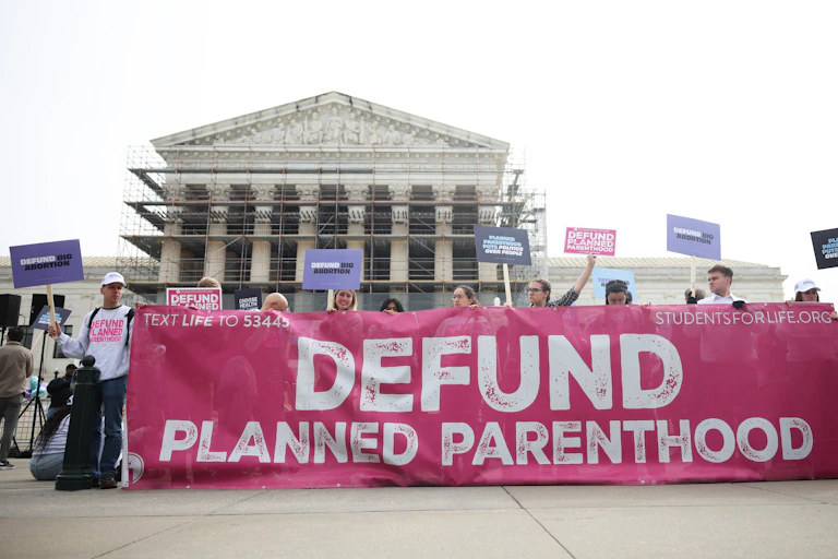 People hold a gigantic pink banner that says 'defund Planned Parenthood' in white letters, with the Supreme Court in the background.