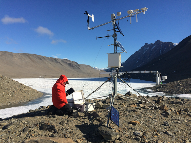 A man in a red jacket sits on the ground and reads a tablet near a metal weather detector.