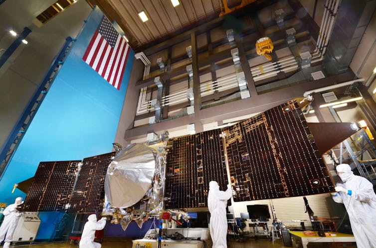 People in white suits stand in front of large metal solar arrays to test them for a space mission to Mars.