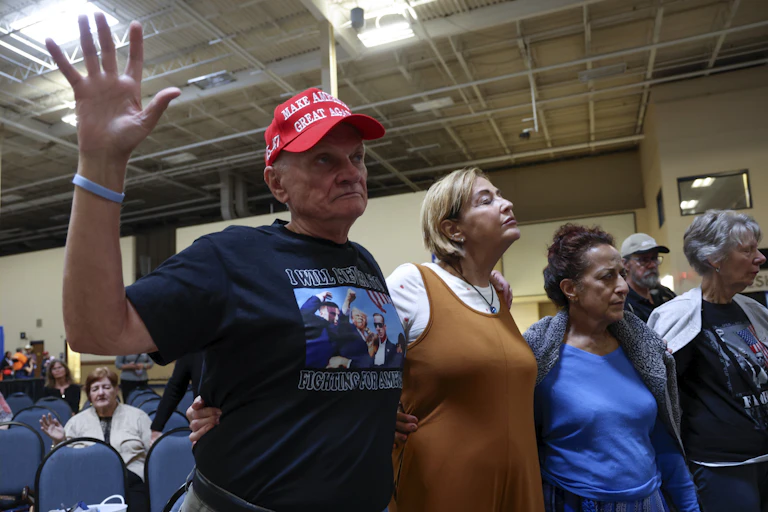 Three individuals stand in prayer with their eyes closed, while several others sit nearby. One of the standing individuals is wearing a MAGA hat
