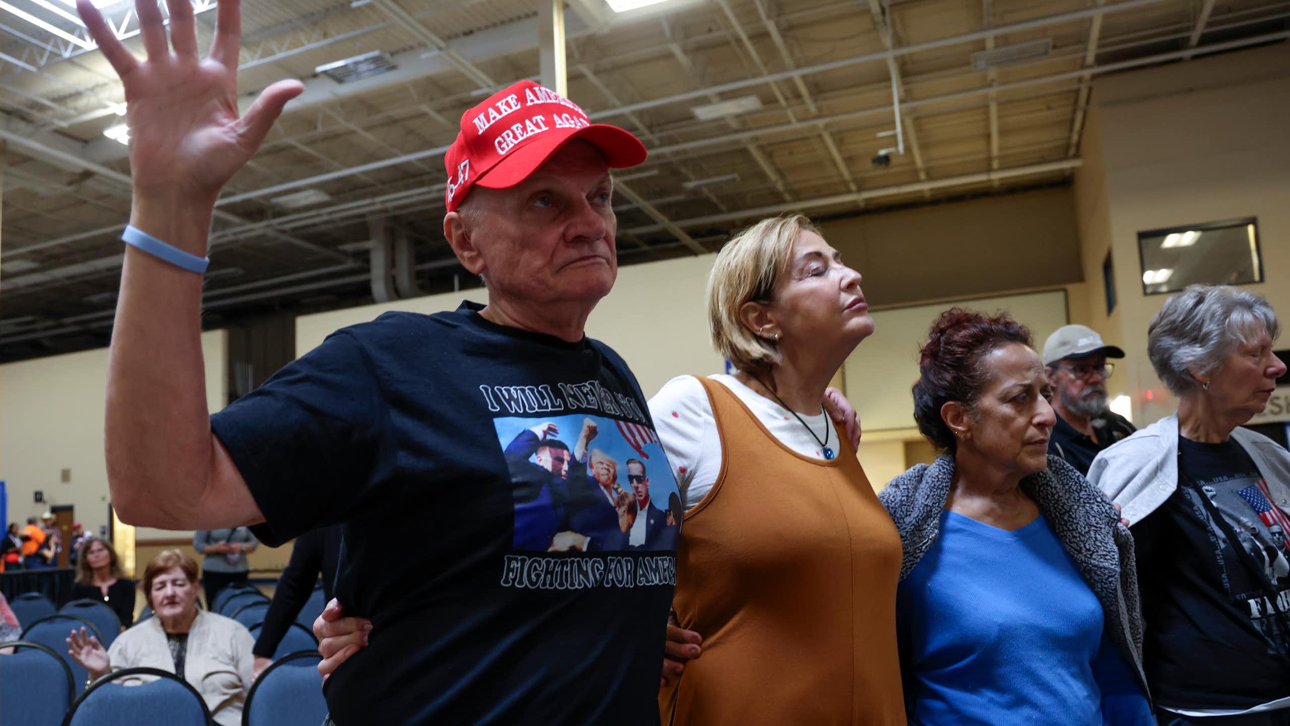 Three individuals stand in prayer with their eyes closed, while several others sit nearby. One of the standing individuals is wearing a MAGA hat