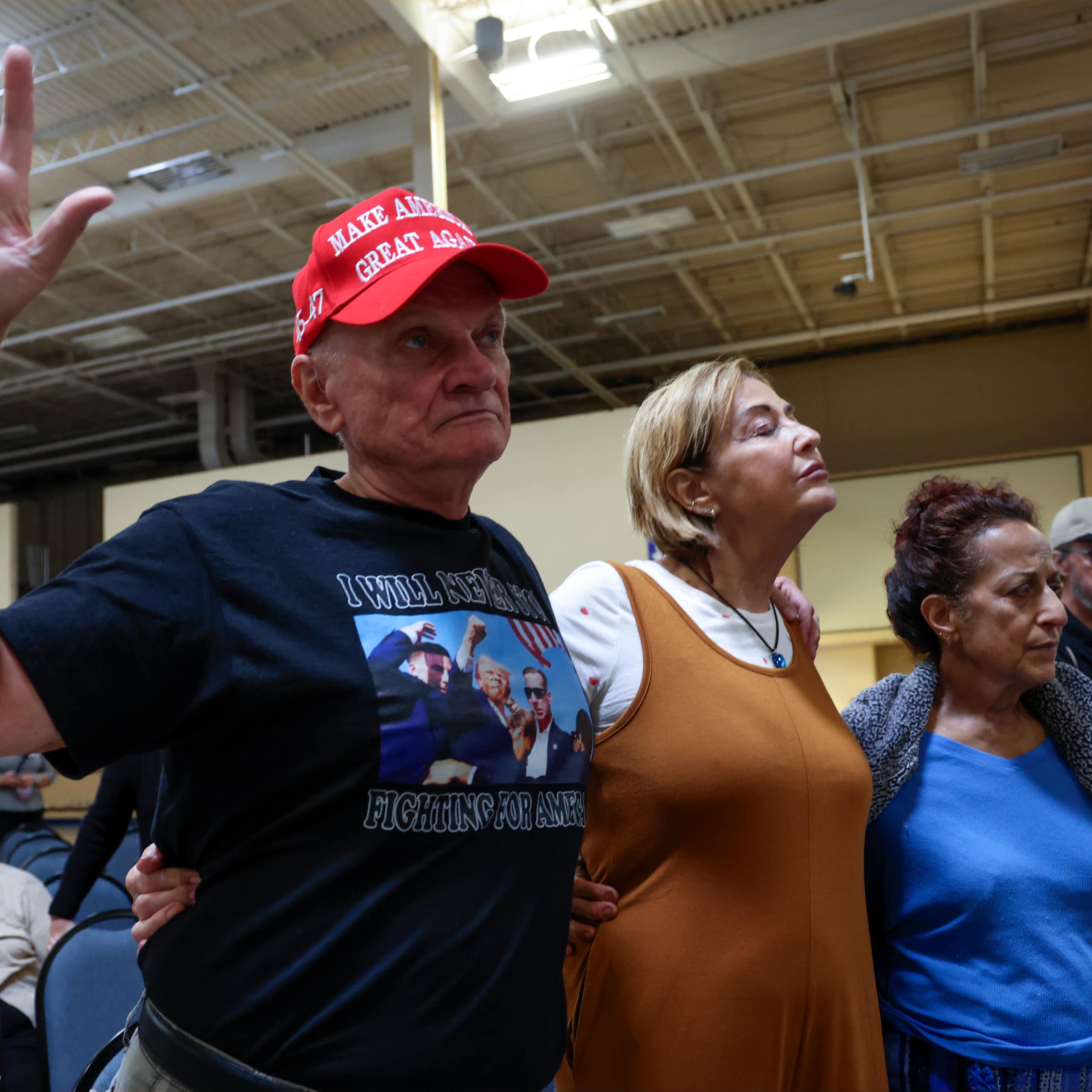 Three individuals stand in prayer with their eyes closed, while several others sit nearby. One of the standing individuals is wearing a MAGA hat