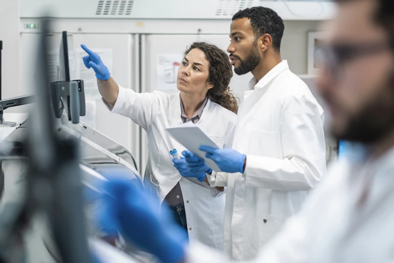 people in white lab coats and blue gloves look at a monitor