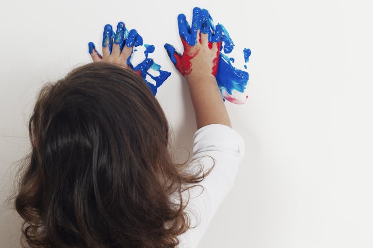 Little girl seen from behind smears blue paint onto a white poster with her hands.
